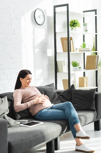 Mujer embarazada feliz sentado en el sofá con la tableta digital y el teléfono inteligente - foto de stock