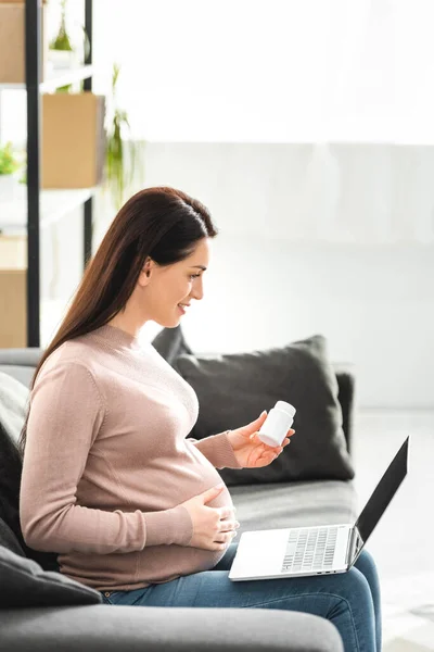 Беременная женщина, держащая таблетки и имеющие онлайн консультации с врачом на ноутбуке дома — стоковое фото