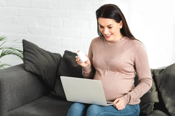 Donna incinta sorridente in possesso di pillole e avendo consultazione online con il medico sul computer portatile a casa — Foto stock