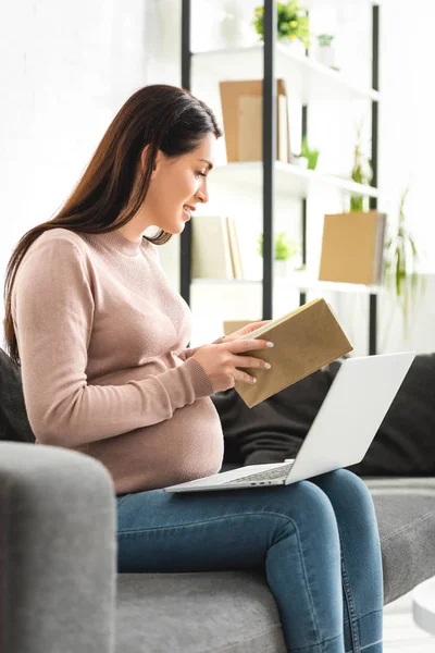 Pregnant woman holding book while having online consultation with doctor on laptop at home — Stock Photo