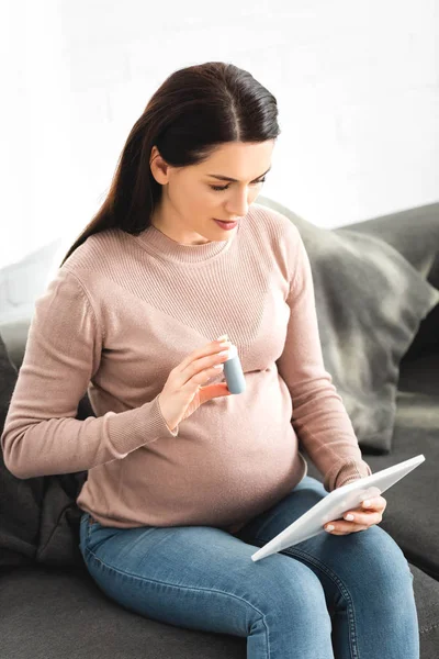 Hermosa mujer embarazada sosteniendo pastillas mientras tiene consulta en línea con el médico en la tableta digital en casa - foto de stock