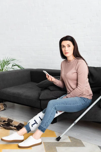 Mujer con la pierna rota y muletas tener consulta en línea con el médico en el teléfono inteligente en el sofá - foto de stock