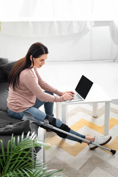 Mulher bonita com perna quebrada e muletas tendo consulta on-line com o médico no laptop em casa — Fotografia de Stock