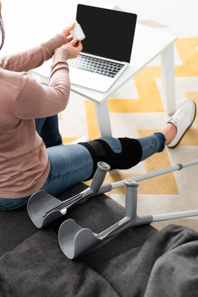 Abgeschnittene Ansicht einer Frau mit gebrochenem Bein und Krücken, die Tabletten in der Hand hält und am Laptop online mit dem Arzt Rücksprache hält — Stockfoto