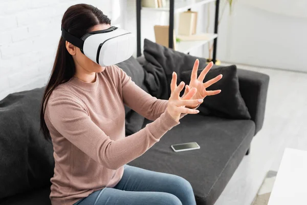 Mujer emocional usando auriculares de realidad virtual mientras está sentado en el sofá con teléfono inteligente - foto de stock