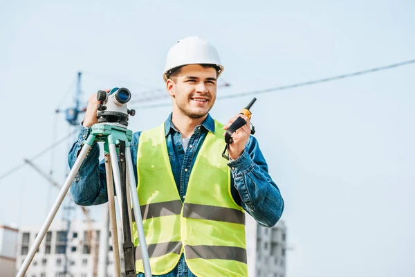 Arpenteur souriant avec niveau numérique parlant sur le poste de radio avec chantier en arrière-plan — Photo de stock