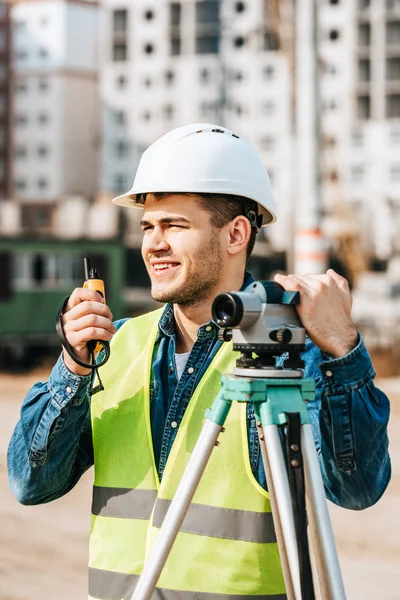 Smiling surveyor with digital level talking on radio set with construction site on background — Stock Photo