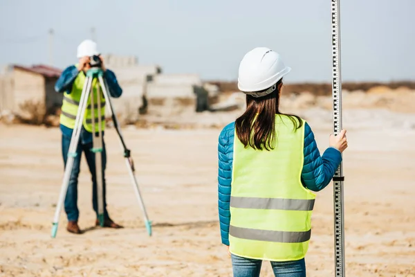 Selective focus of surveyor holding ruler and colleague with digital level on background — Stock Photo