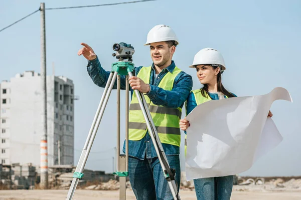 Vermessungsingenieur mit digitaler Ebene zeigt mit Blaupause auf Kollegen — Stockfoto