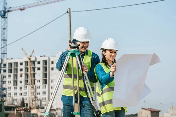 Smiling surveyors with digital level looking at blueprint on construction site — Stock Photo