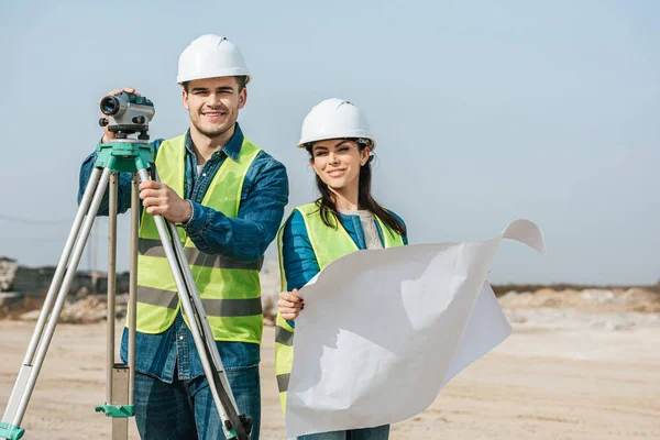 Smiling surveyors with digital level and blueprint looking at camera — Stock Photo