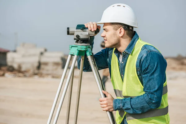 Vue latérale de l'arpenteur en casque dur regardant tout au long du niveau numérique sur le chantier de construction — Photo de stock