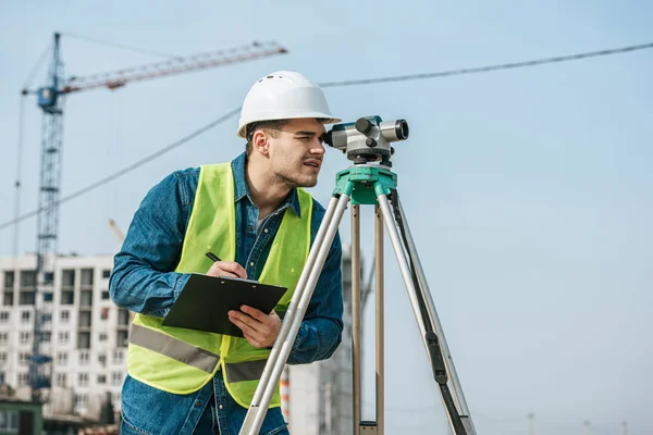 Surveyor regardant à travers le niveau numérique et l'écriture sur presse-papiers — Photo de stock