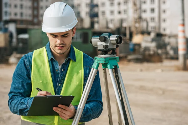 Surveyor escrevendo na área de transferência ao lado do nível digital no canteiro de obras — Fotografia de Stock