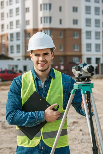 Agrimensor sujetando el portapapeles y el nivel digital y sonriendo a la cámara en el sitio de construcción - foto de stock