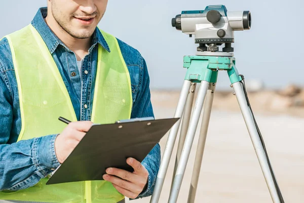 Cropped shot of smiling surveyor writing on clipboard beside digital level — Stock Photo