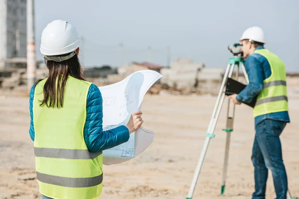 Selective focus of surveyor with blueprint and colleague using digital level on construction site — Stock Photo