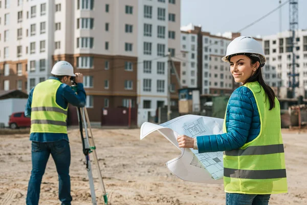 Enfoque selectivo del topógrafo con el plano sonriendo a la cámara y colega utilizando el nivel digital en el sitio de construcción - foto de stock