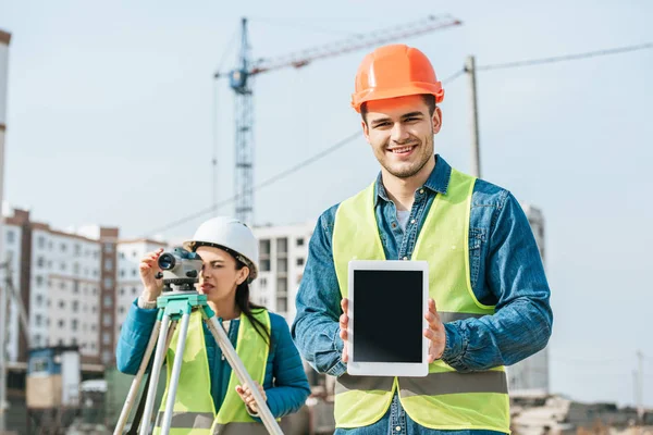 Sorridente geometra con tablet digitale con schermo bianco e collega con livello di misurazione in background — Foto stock