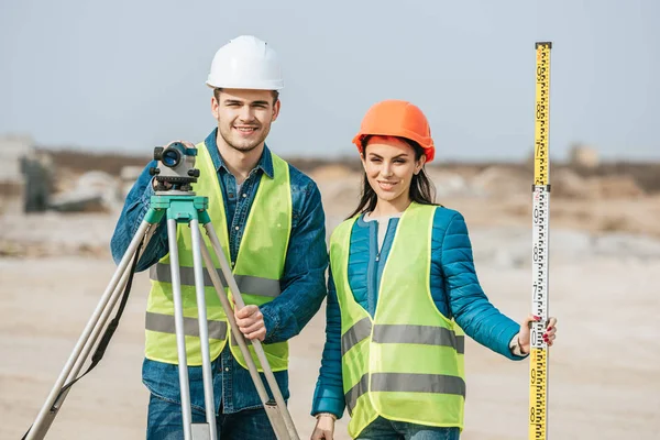 Lächelnde Vermessungsingenieure mit Lineal und digitaler Ebene schauen in die Kamera — Stockfoto