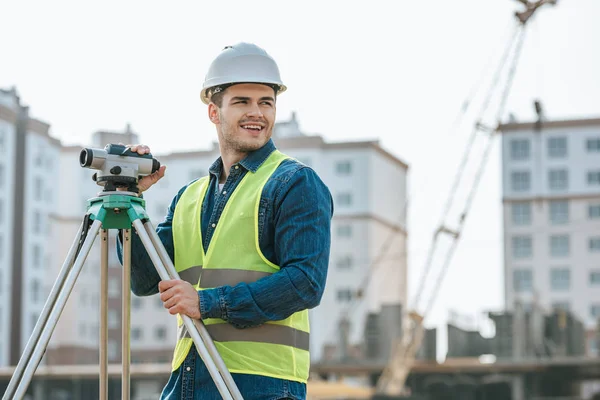 Sonriente topógrafo con nivel digital mirando hacia otro lado en el sitio de construcción - foto de stock