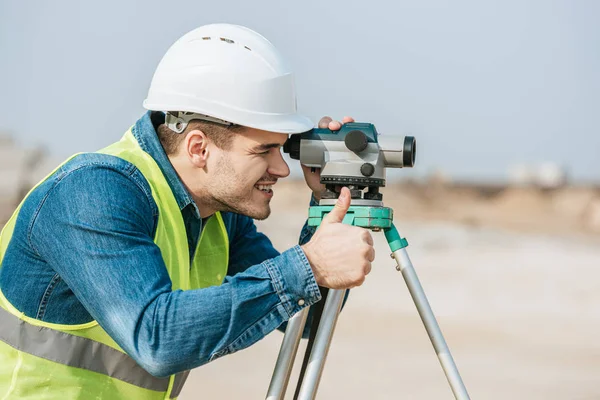 Lächelnder Vermessungsingenieur blickt durch digitale Ebene und zeigt Daumen-hoch-Geste — Stockfoto