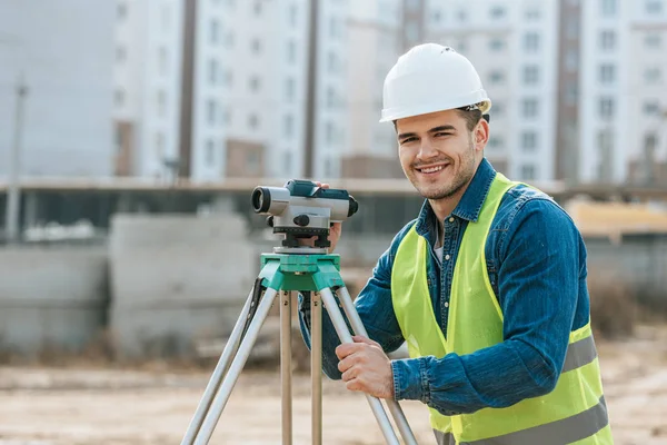 Lächelnder Vermessungsingenieur mit digitaler Ebene blickt auf die Kamera auf der Baustelle — Stockfoto