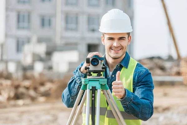 Surveyor con livello digitale sorridente alla fotocamera e mostrando il pollice verso l'alto gesto — Foto stock