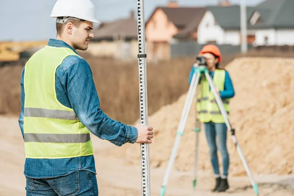 Selective focus of surveyor with ruler and colleague with digital level on background — Stock Photo