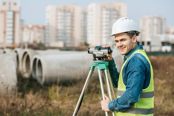 Surveyor avec niveau numérique souriant à la caméra avec des matériaux de construction en arrière-plan — Photo de stock