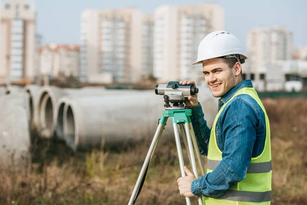 Agrimensor con nivel digital sonriendo a la cámara con materiales de construcción al fondo - foto de stock