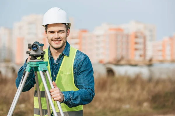 Smiling surveyor in hardhat and high visibility jacket holding digital level — Stock Photo
