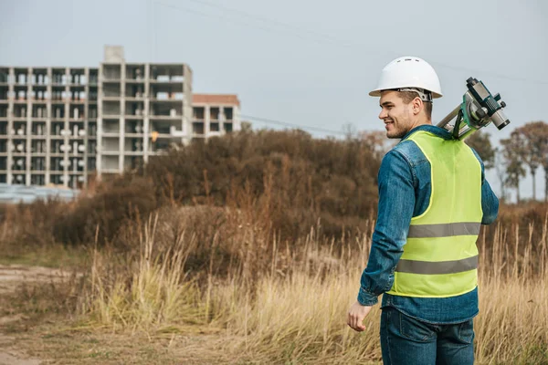 Surveyor holding digital level with field and construction site at background — Stock Photo