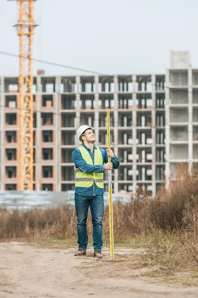 Lächelnder Vermessungsingenieur hält Lineal mit Baustelle im Hintergrund — Stockfoto