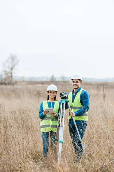 Agrimensores con nivel digital y tableta sonriendo a cámara en campo - foto de stock