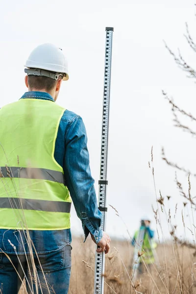 Selective focus of surveyor with ruler and colleague with digital level in field — Stock Photo