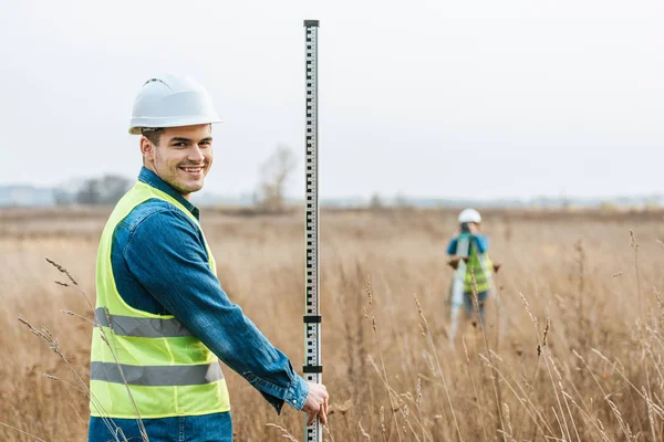 Selective focus of smiling surveyor with ruler and colleague with digital level in field — Stock Photo