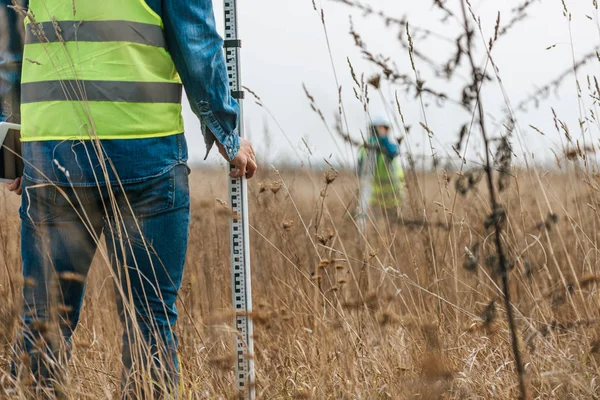 Selective focus of surveyor with ruler and colleague with digital level in field — Stock Photo