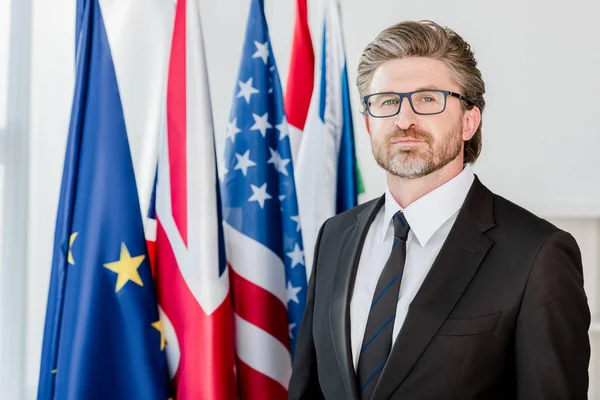 Schöner Diplomat mit Brille, der in die Kamera neben Flaggen blickt — Stockfoto