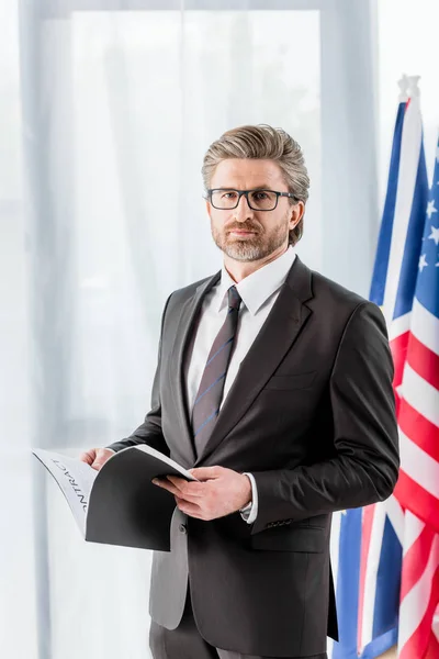 Guapo diplomático en gafas celebración carpeta cerca american bandera - foto de stock