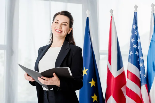 Happy diplomat in formal wear holding folder near flags — Stock Photo