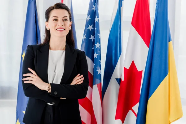 Feliz diplomático de pie con los brazos cruzados cerca de banderas - foto de stock