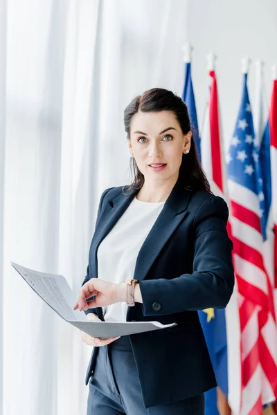 Attractive diplomat holding folder and looking at camera near flags — Stock Photo