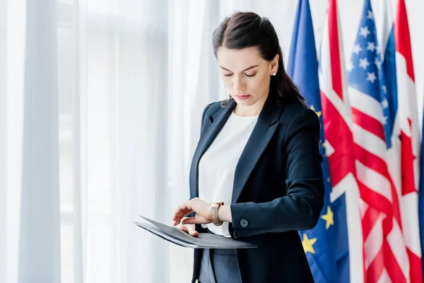 Attractive diplomat holding folder and looking at watch near flags in embassy — Stock Photo