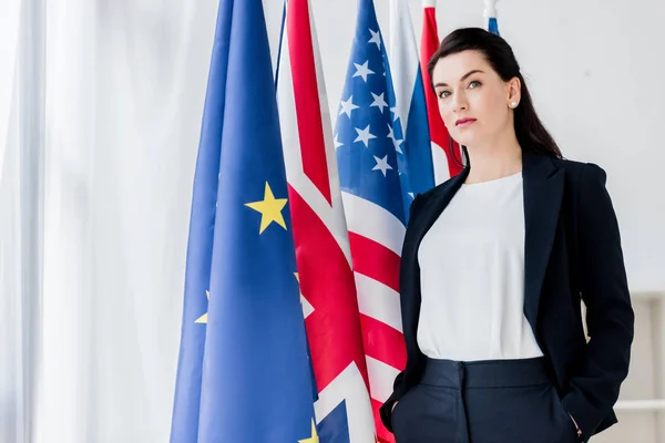 Beautiful diplomat standing with hands in pockets near flags and looking at camera — Stock Photo