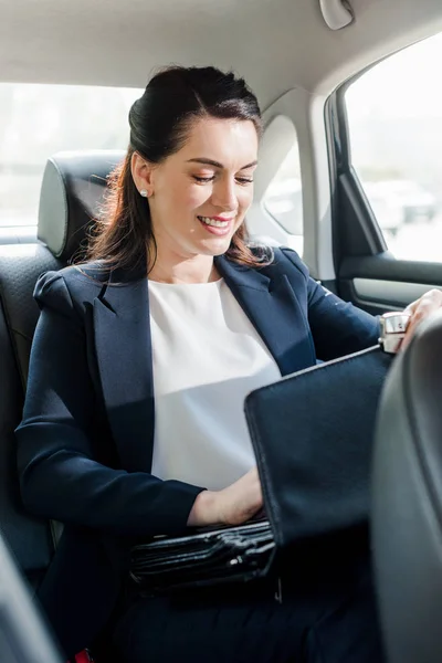 Embaixador atraente sorrindo enquanto sentado no carro e tocando pasta — Fotografia de Stock