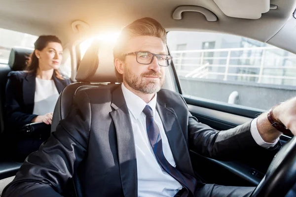 Fröhlicher Diplomat fährt Auto und blickt in Kamera neben Frau — Stockfoto