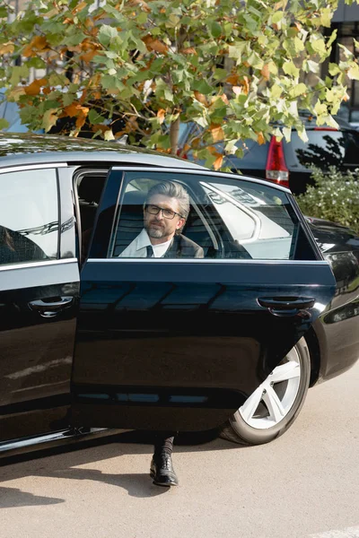 Beau diplomate dans des lunettes assis dans la voiture avec porte de voiture ouverte — Photo de stock