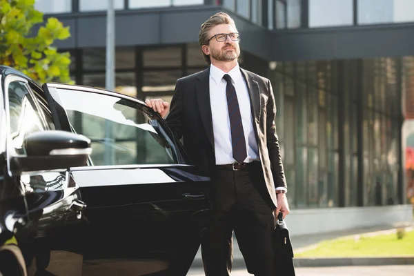 Schöner Diplomat mit Brille steht mit Aktentasche neben schwarzem Auto — Stockfoto