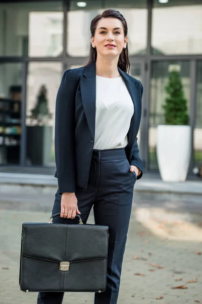 Beautiful diplomat holding briefcase and standing with hand in pocket outside — Stock Photo